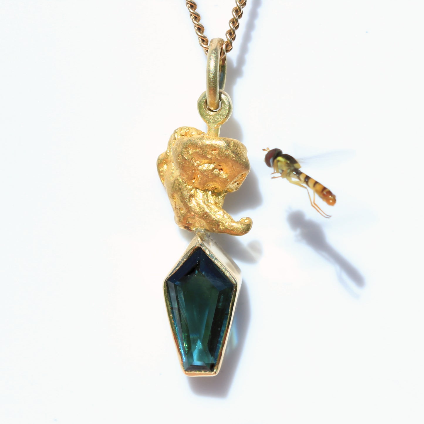 Close-up of Australian parti sapphire kite-cut pendant topped with a natural gold nugget; small hoverfly nearby on white background.