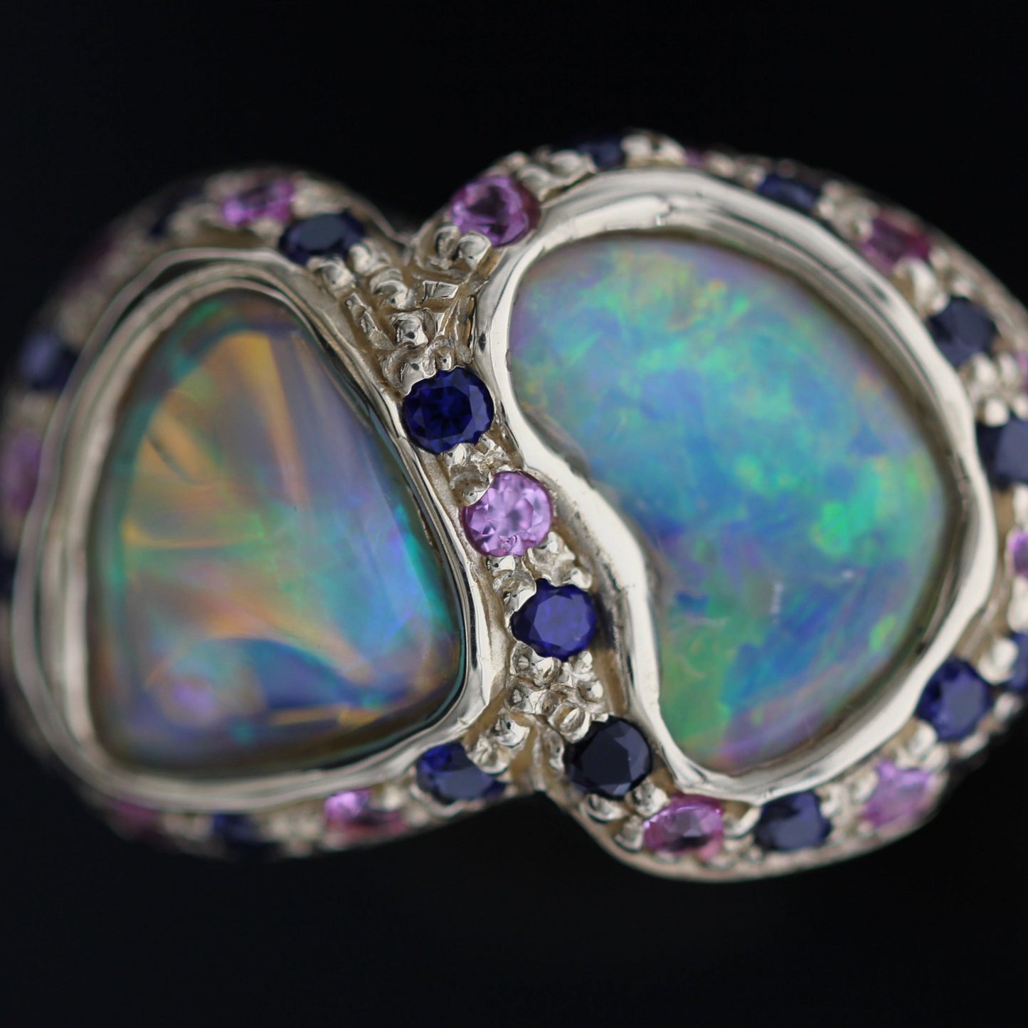 Macro detail of twin opals surrounded by alternating blue and purple sapphires in a textured 14ct yellow gold mount.