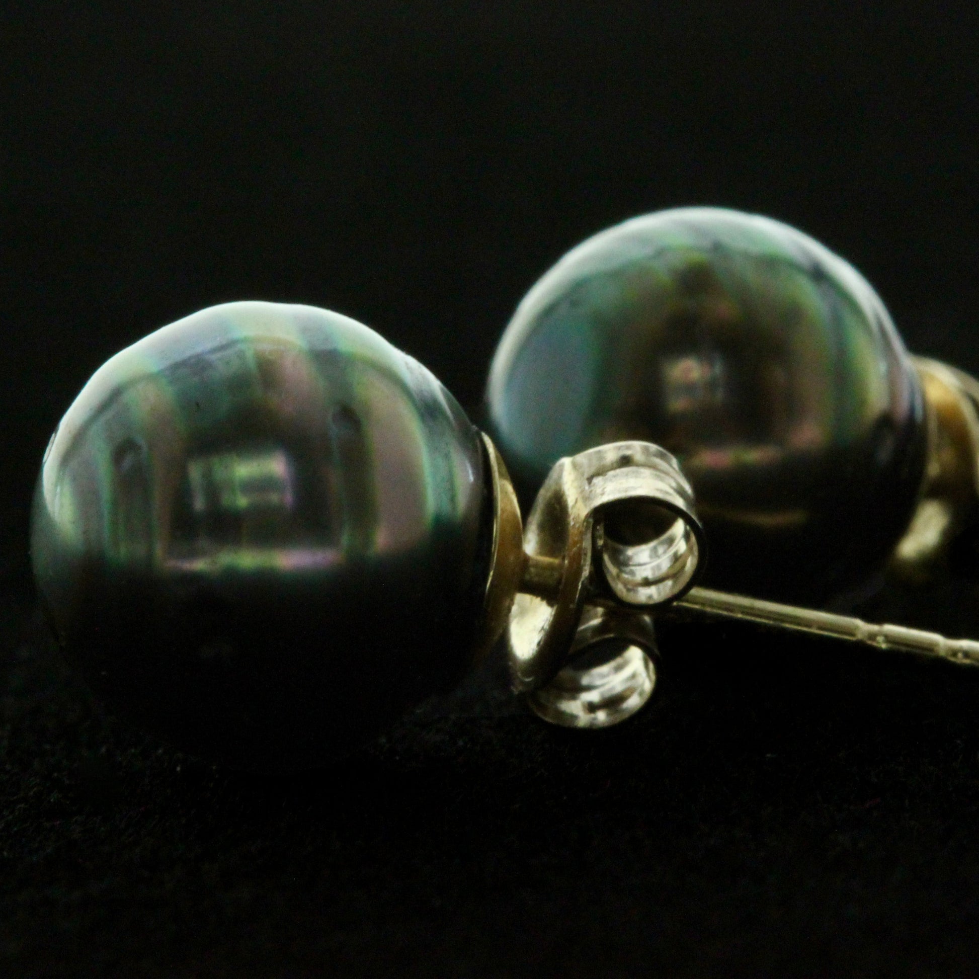 Macro view of 10mm black Tahitian pearl stud earrings with 14ct yellow gold posts and scrolls on a dark background.