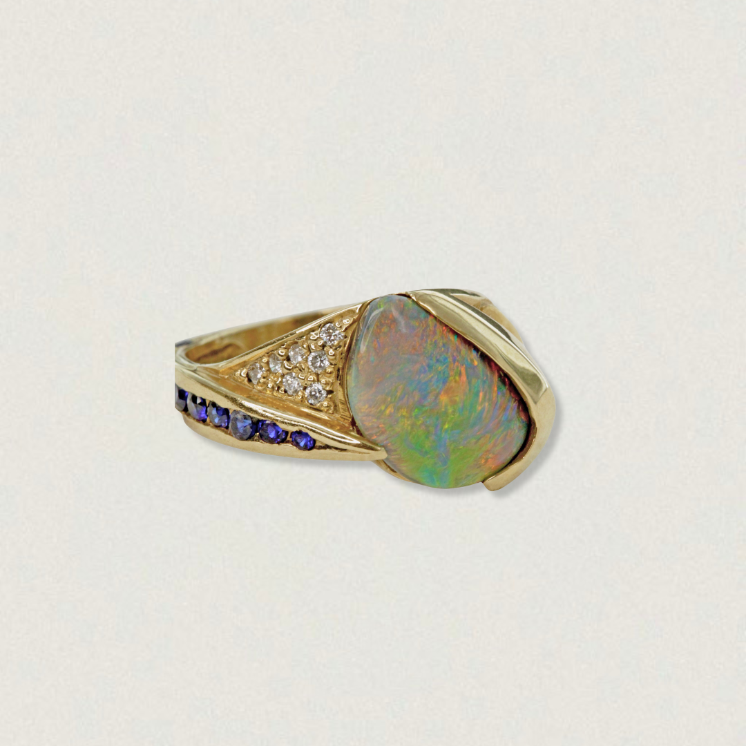 2.29ct Lightning Ridge black opal statement ring with blue sapphires and diamond accents in 18ct yellow gold, angled product view on light background.