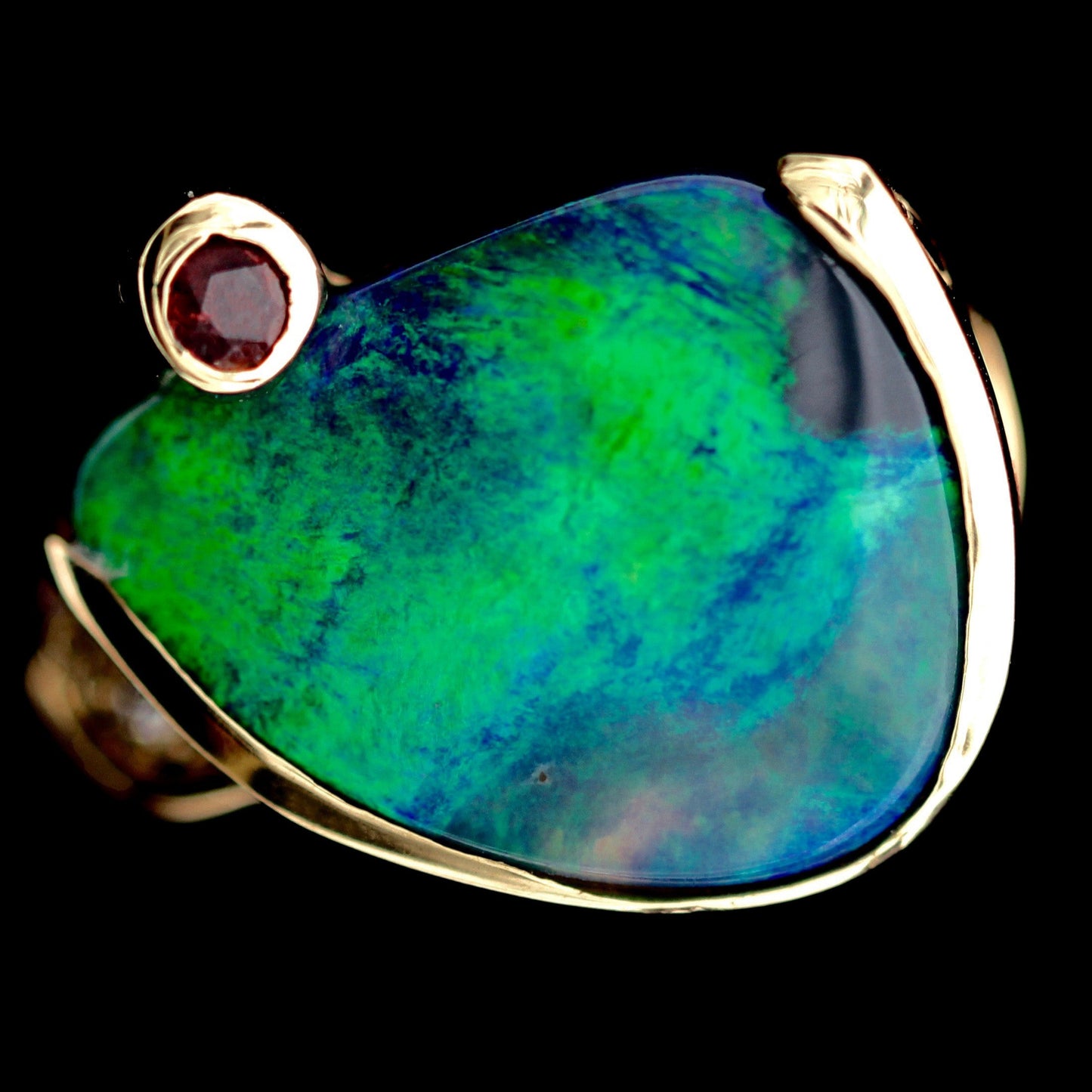 Close-up of freeform green-blue Lightning Ridge opal with round ruby accent in 18ct yellow gold setting.