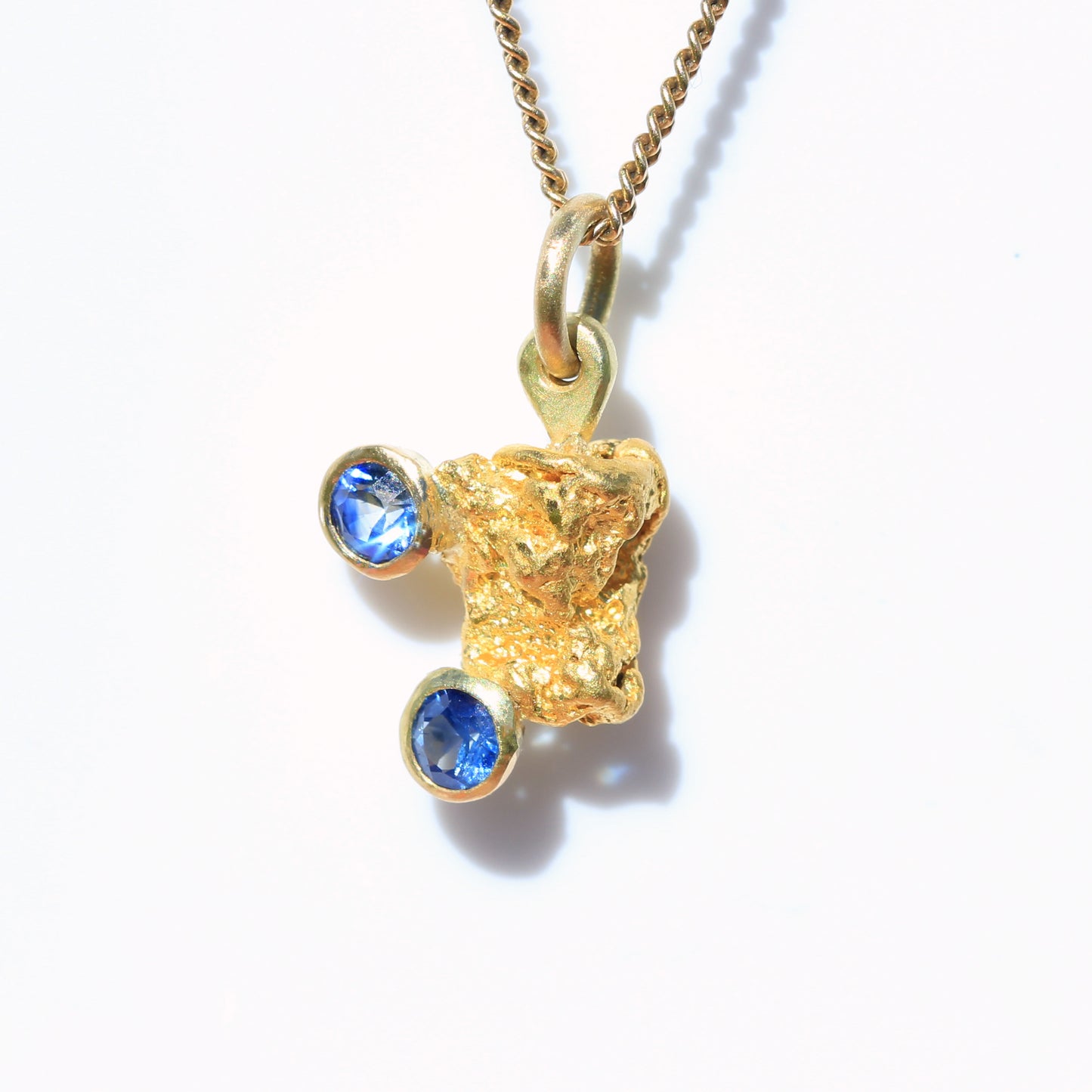 Close-up of natural Australian gold nugget pendant set with two bright blue parti sapphires on chain, front view on white.