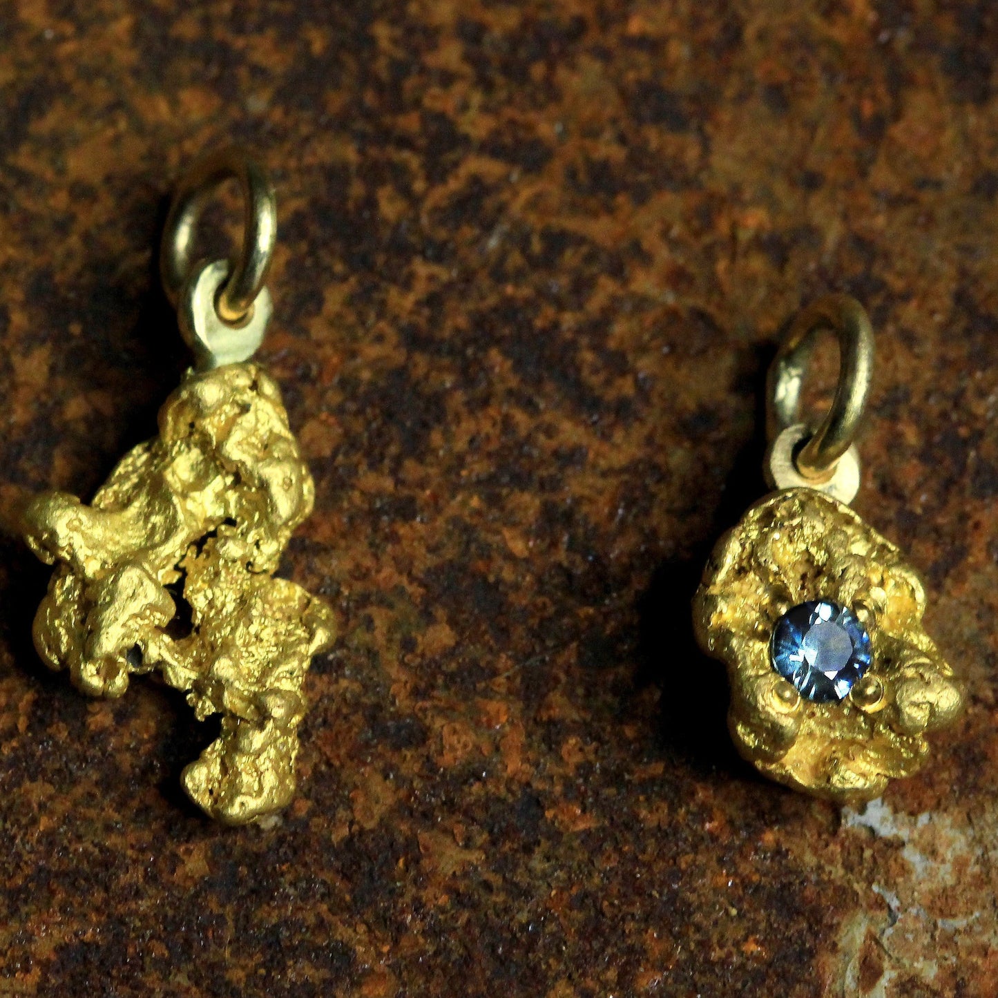 Pair of Australian natural gold nugget pendants—one plain and one set with a round blue parti sapphire—on a textured stone background