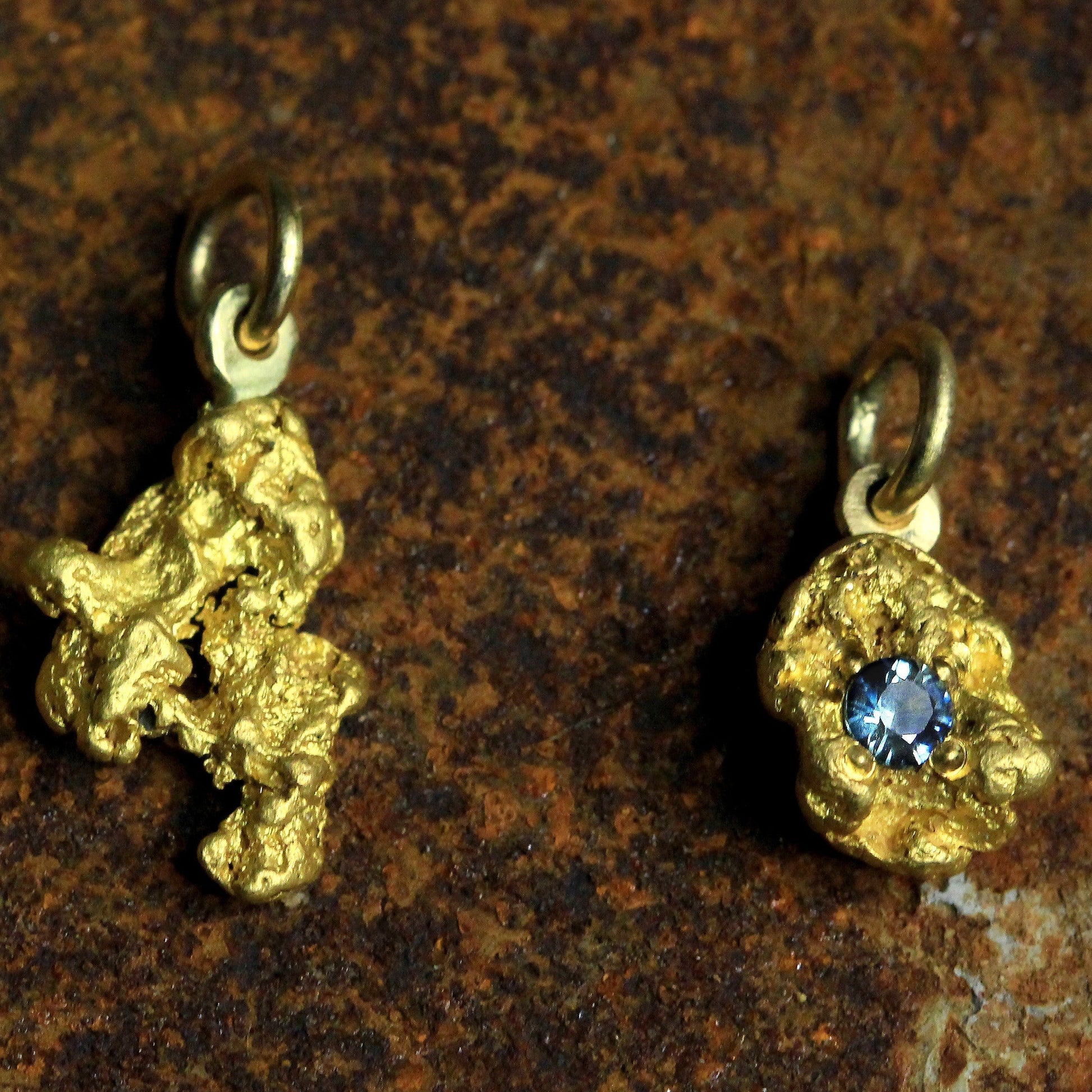Pair of Australian natural gold nugget pendants—one plain and one set with a round blue parti sapphire—on a textured stone background