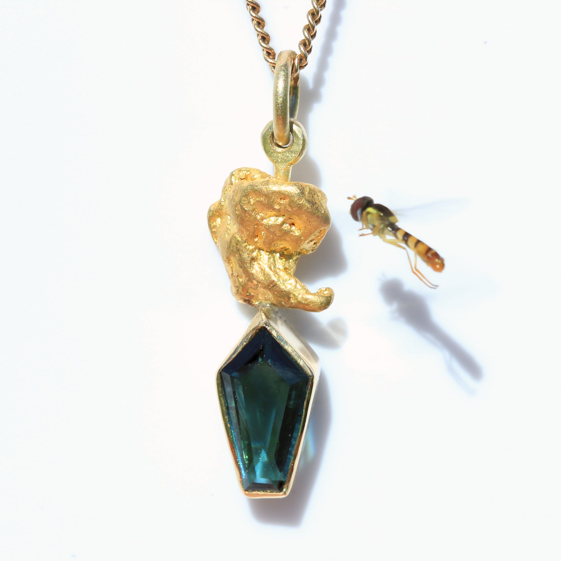 Close-up of Australian parti sapphire kite-cut pendant topped with a natural gold nugget; small hoverfly nearby on white background.