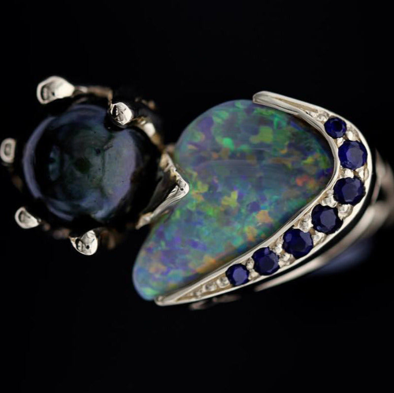 Close-up of freeform Lightning Ridge opal with blue sapphire pavé on 14ct yellow gold ring, next to black Tahitian pearl