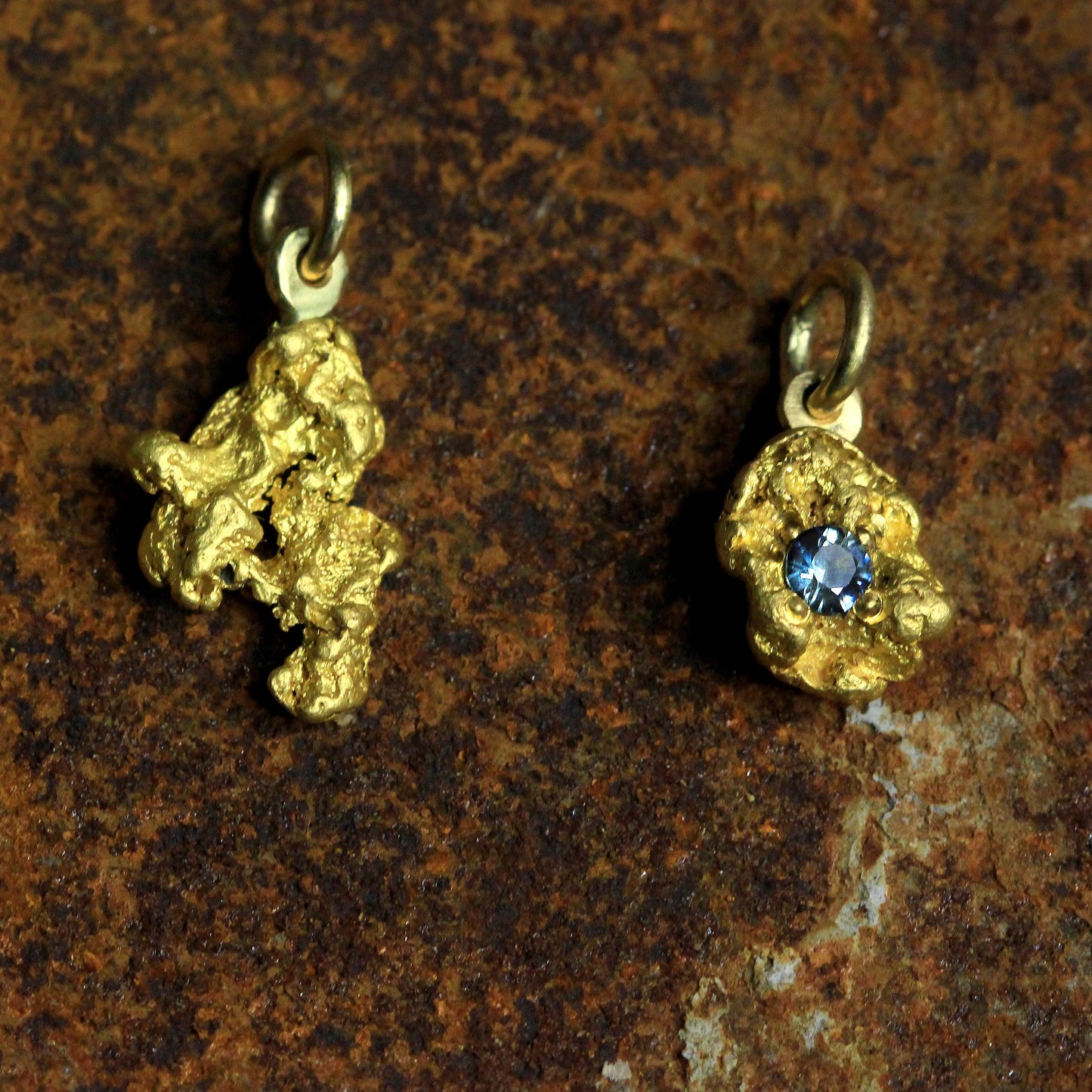 Two natural gold nugget pendants side by side; one set with a round blue sapphire, on textured stone background.