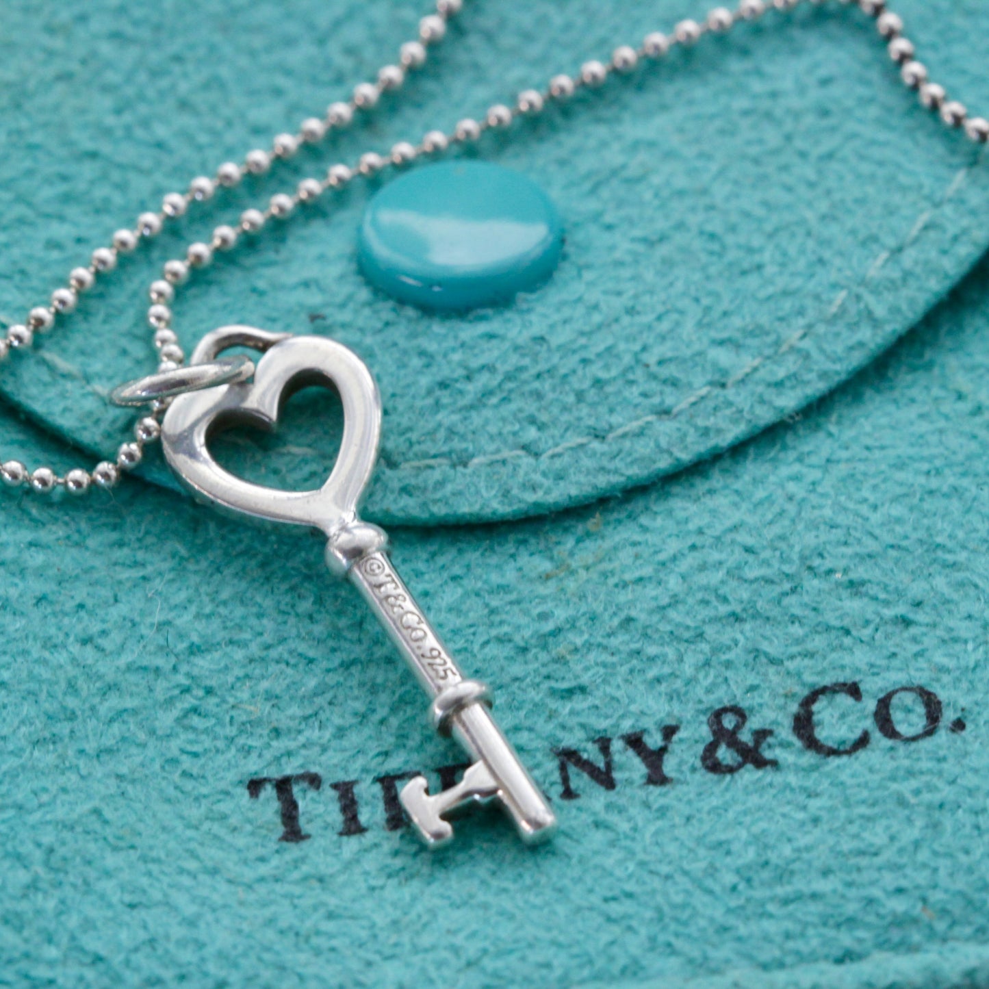 Close-up of Tiffany & Co. sterling heart key charm on bead chain over Tiffany blue pouch.