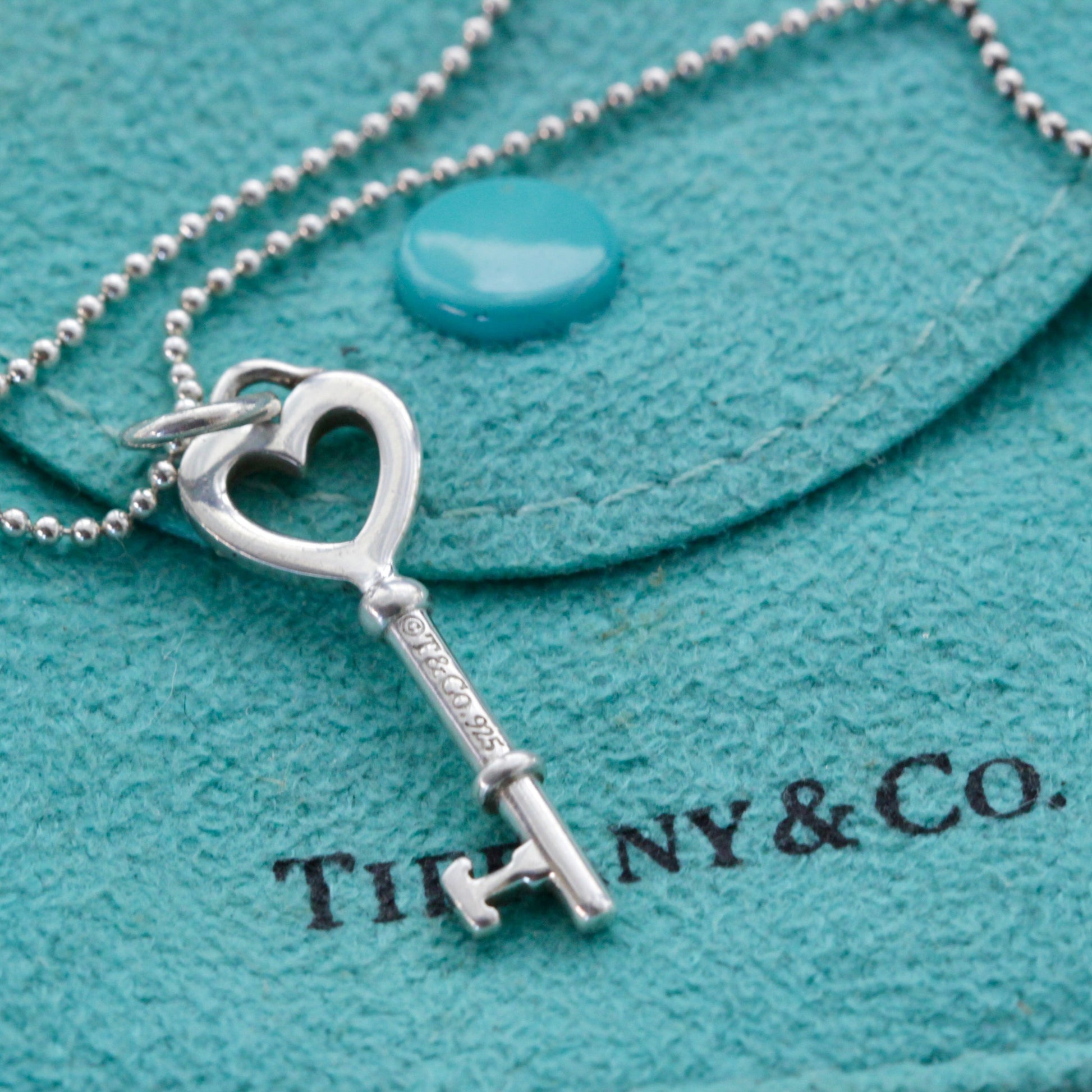 Close-up of Tiffany & Co. sterling heart key charm on bead chain over Tiffany blue pouch.